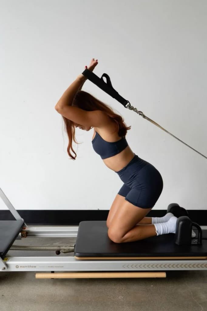 A woman kneeling on a Pilates reformer, pulling straps overhead, in a bright studio.